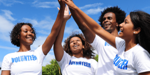 Three females and one male, of various ethnicities, raise hands together as each person wears a shirt with the label volunteer