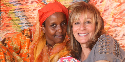 Black woman in orange headband smiles cheek-to-cheek at camera next to white woman with blond hair